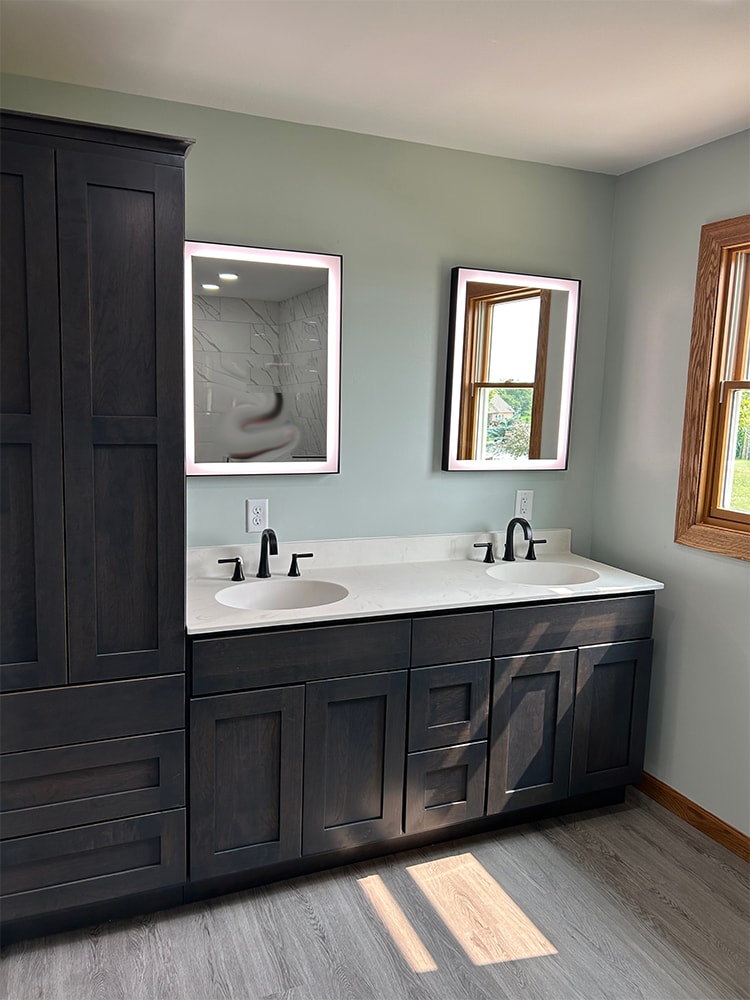 Siding, bathroom, garage & 1/2 bath — after: completed master bath double vanity with dark cabinetry, white countertop, and lighted mirrors — Attica, Ohio