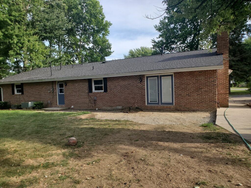 Deck addition Findlay — before: front of brick ranch home with gravel ground, no deck, blue entry door, and mature trees, Findlay, Ohio