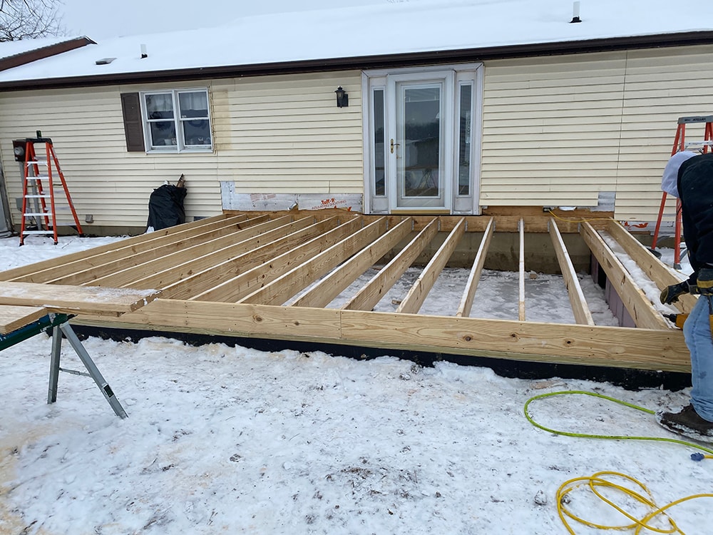 Room addition Fostoria — framing stage: floor joist system laid on foundation in winter snow with crew working and ladders staged, Fostoria, Ohio