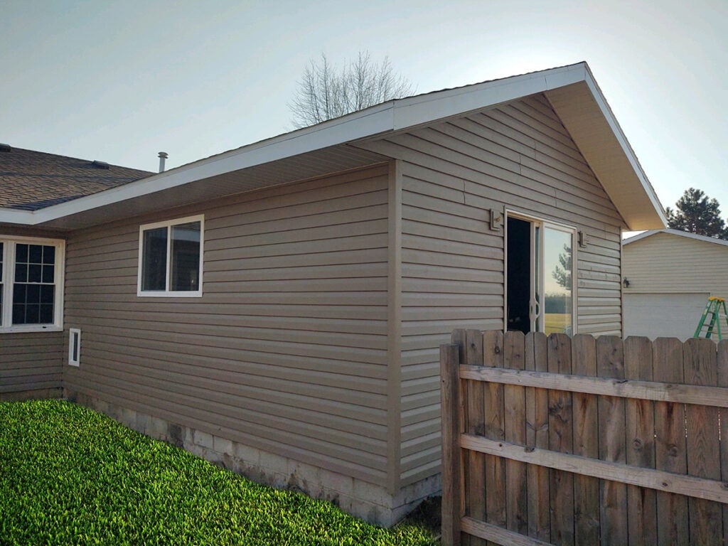 Large room addition Findlay — after: completed exterior side view with new tan vinyl siding, gable roof, sliding glass door, and green lawn at dusk, Findlay, Ohio