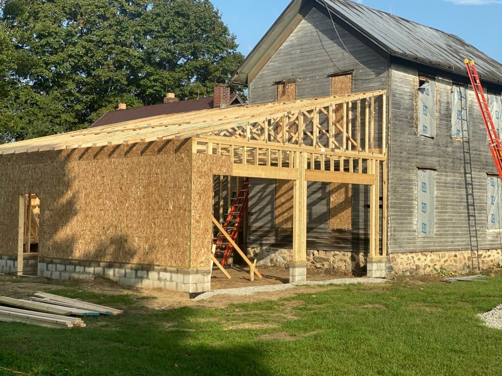 Metal roof large addition Gibsonburg — during: large addition framed with OSB sheathing on block foundation, open bay framing and orange ladder visible, original home in background, Gibsonburg, Ohio