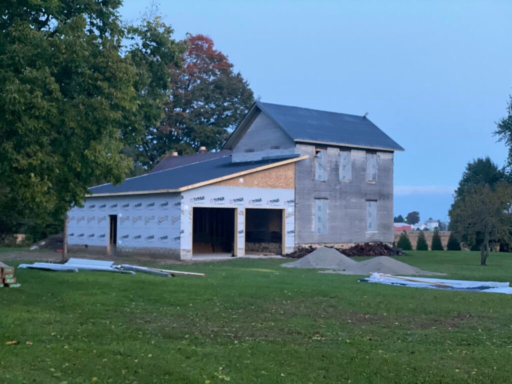 Metal roof large addition Gibsonburg — during: wide addition with Typar house wrap installed, new metal roof on addition, open garage bays, gravel and materials staged on lawn, dusk light, Gibsonburg, Ohio