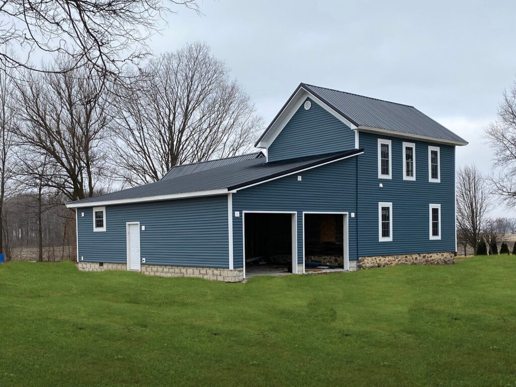 Metal roof large addition Gibsonburg — after: rear of completed home showing large addition with blue vinyl siding, metal roof, open garage bay, white trim, overcast sky, Gibsonburg, Ohio
