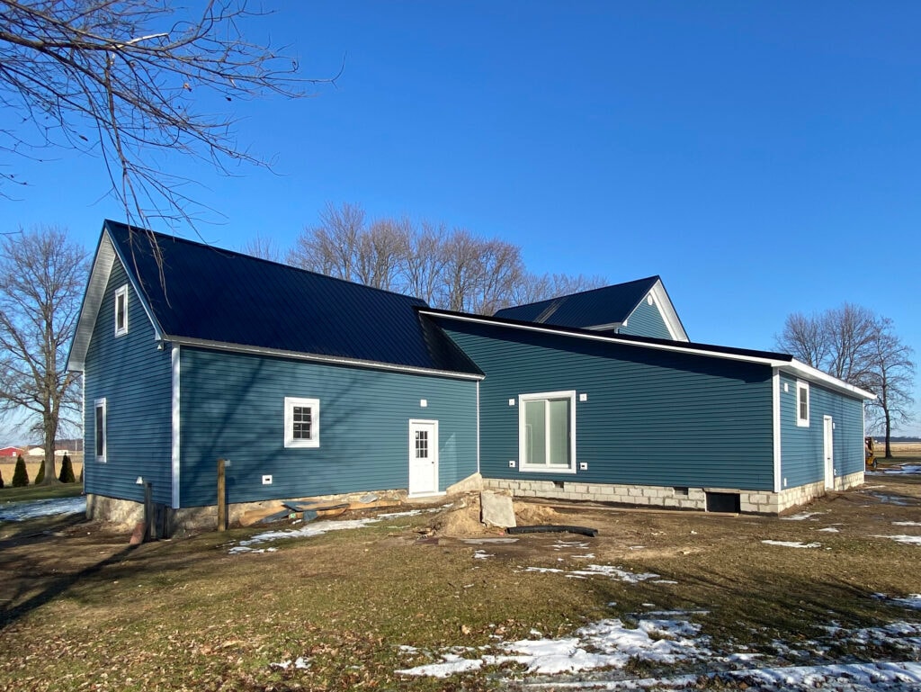 Metal roof large addition Gibsonburg — after: full rear view of completed home and addition with blue vinyl siding, dark metal roof across all rooflines, white trim, winter sun, Gibsonburg, Ohio
