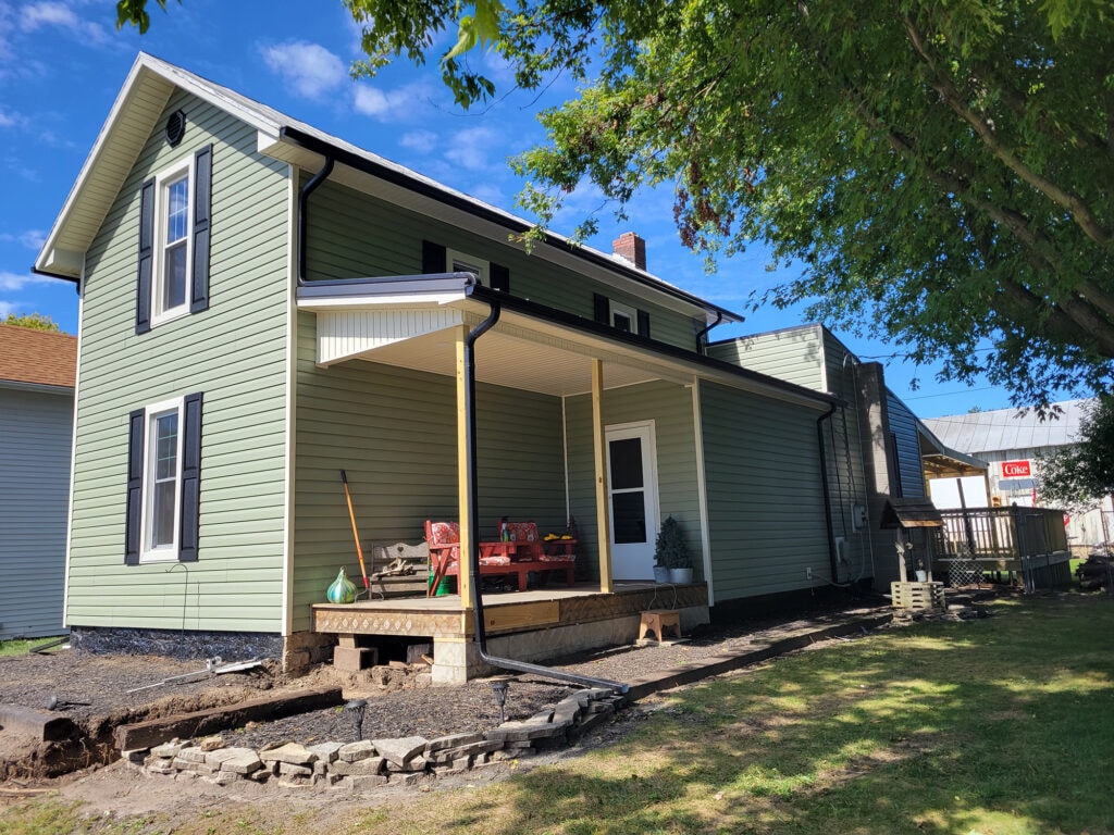 New siding and deck — after, front view with new sage green vinyl siding, black shutters, and new front porch deck, Benton Ridge, Ohio
