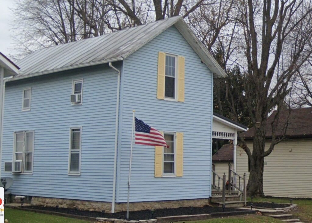 New siding and deck — before, side view of original blue siding with American flag, Benton Ridge, Ohio
