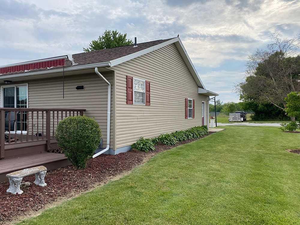 New siding Tiffin — after: side view of home with new tan vinyl siding, red shutters, deck, and stone garden bench, Tiffin, Ohio