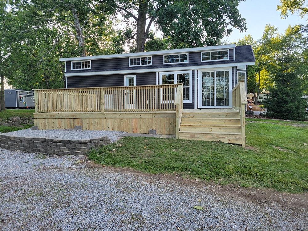 New treated deck — after: dark gray park model with completed full-width treated wood deck and stone fire pit area — Wapakoneta campground