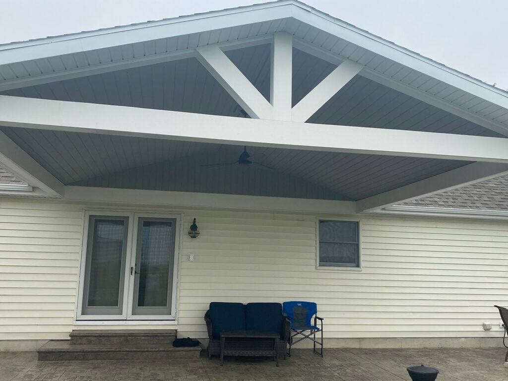 Patio addition Fostoria — after: straight-on exterior view of completed gable roof structure showing full beam work, T&G ceiling, ceiling fan, patio door, and stamped concrete patio with navy chairs, Fostoria, Ohio