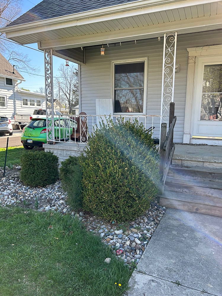 Porch railing replacement — before: side angle view with original ornate iron railing and overgrown bushes — Findlay, Ohio