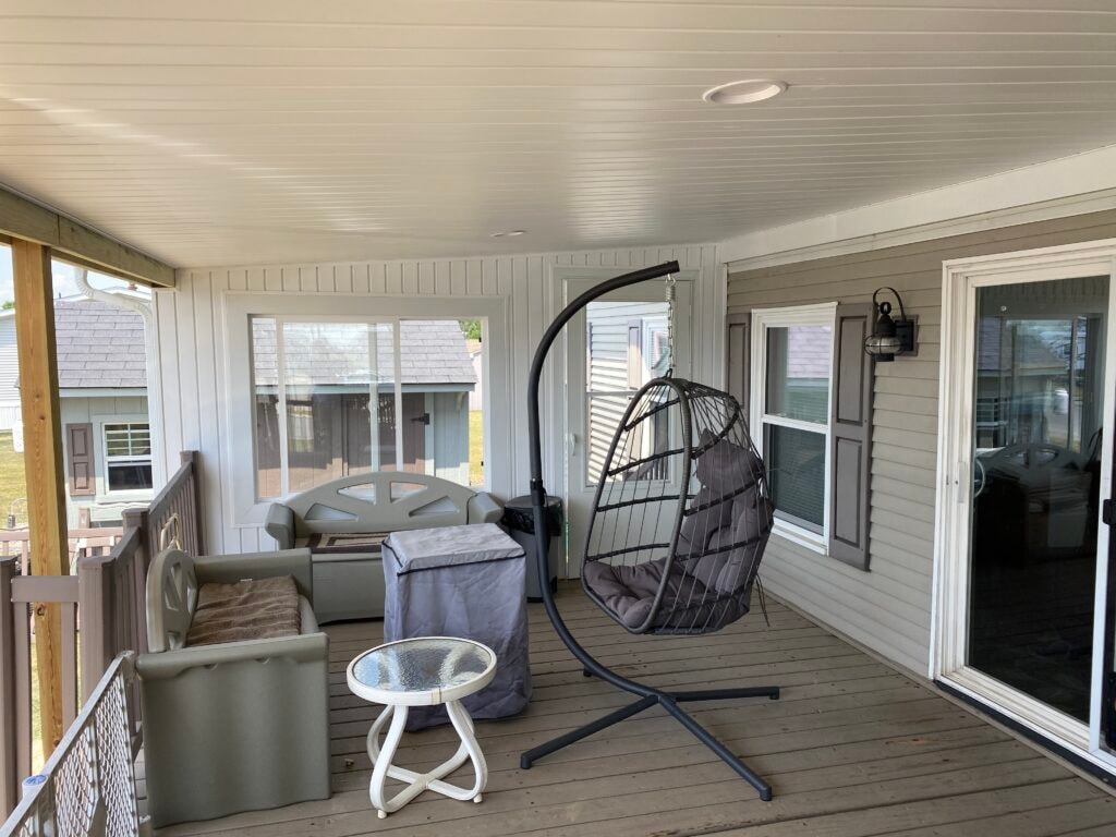 Roof addition existing deck Findlay — after: interior of covered deck with white beadboard ceiling, recessed light, egg chair on stand, gray patio sofa, sliding glass door, and lantern wall sconce, Findlay, Ohio