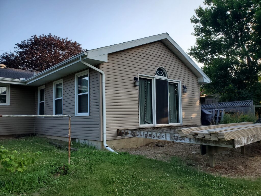 Siding job Findlay — before: rear of home with original tan vinyl siding, old deck framing staged on ground, summer evening light, Findlay, Ohio