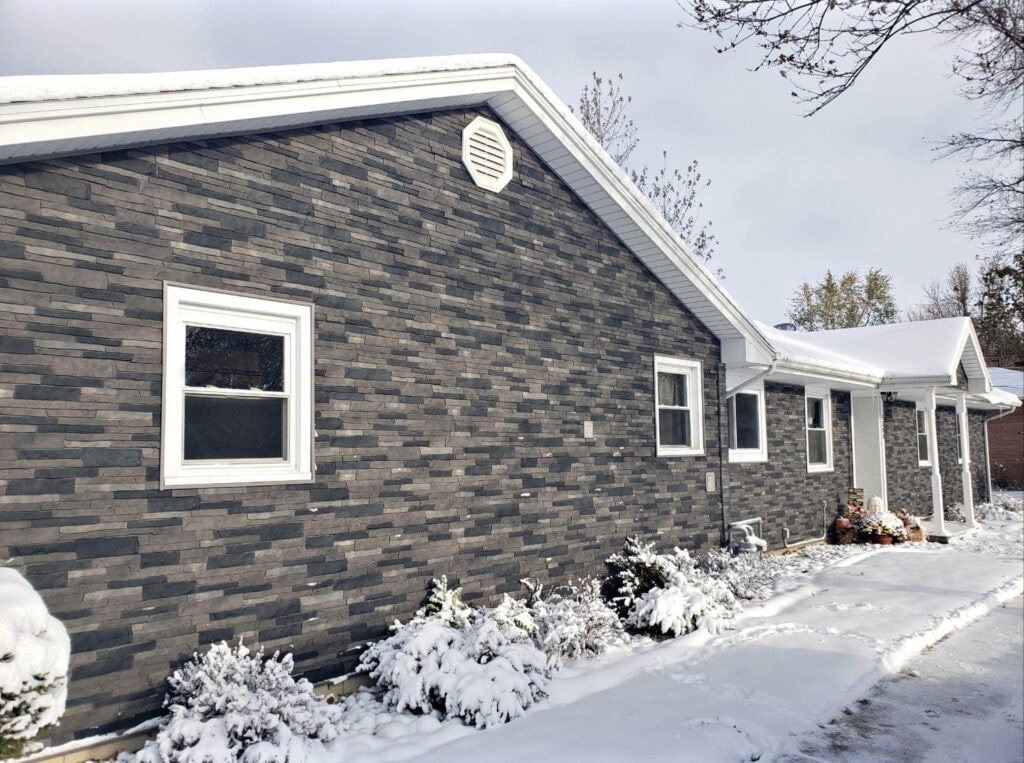 Stone siding Findlay — after: completed exterior in winter snow showing full dark gray stacked stone siding, new white-trimmed windows, white gable vent, and snow-covered shrubs, Findlay, Ohio