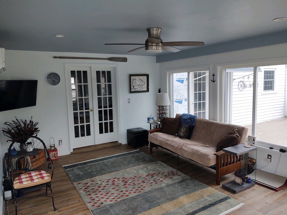 Sunroom transformation Fostoria — after: interior wide angle showing full room with ceiling fan, French doors, futon sofa, area rug, LVP flooring, and large sliding glass door to deck, Fostoria, Ohio
