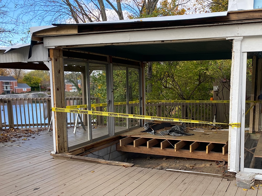 Sunroom transformation Fostoria — demo stage: old sunroom floor removed with caution tape, exposed floor joists, and water view beyond deck, Fostoria, Ohio