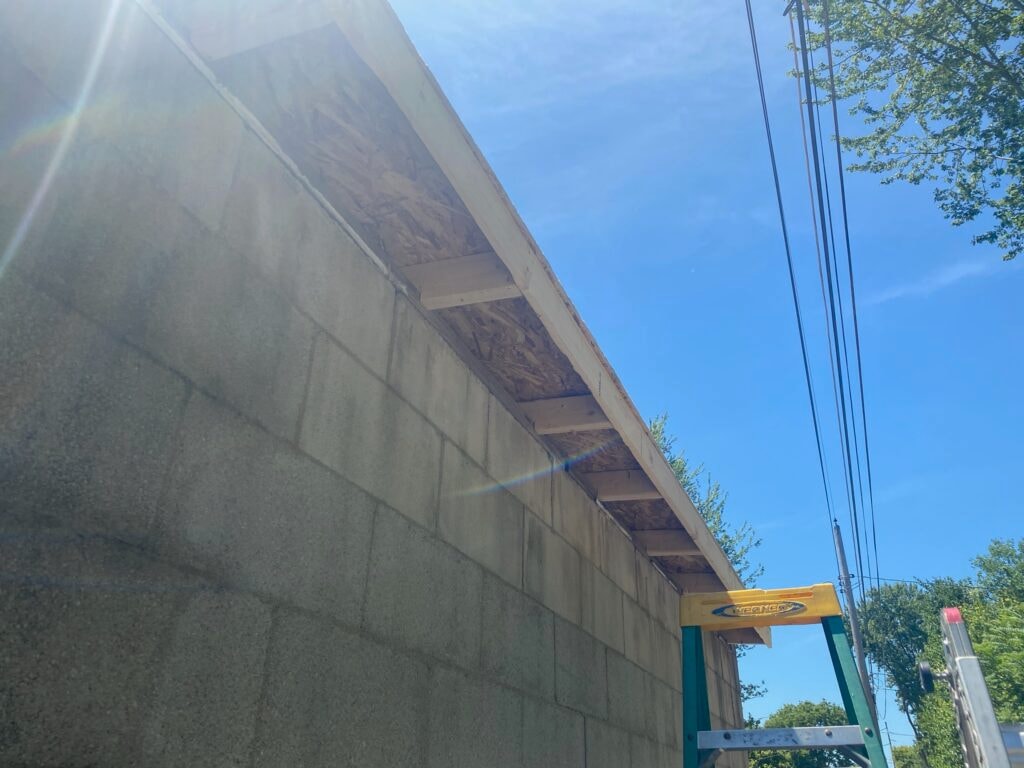 TPO roof replacement Findlay — during: low-angle view of concrete block wall with new fascia board and OSB decking edge installed, ladder visible, blue sky, Findlay, Ohio