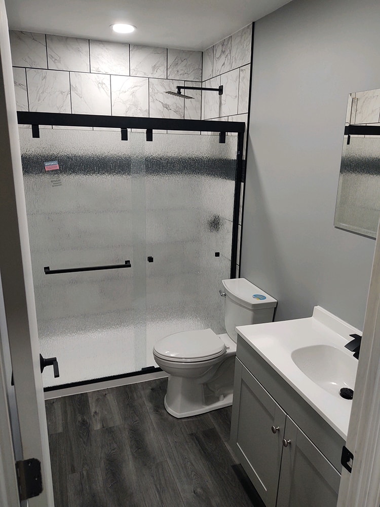 Whole house remodel after — bathroom wide view with marble tile shower, black fixtures, gray vanity, and gray LVP flooring, Fostoria, Ohio