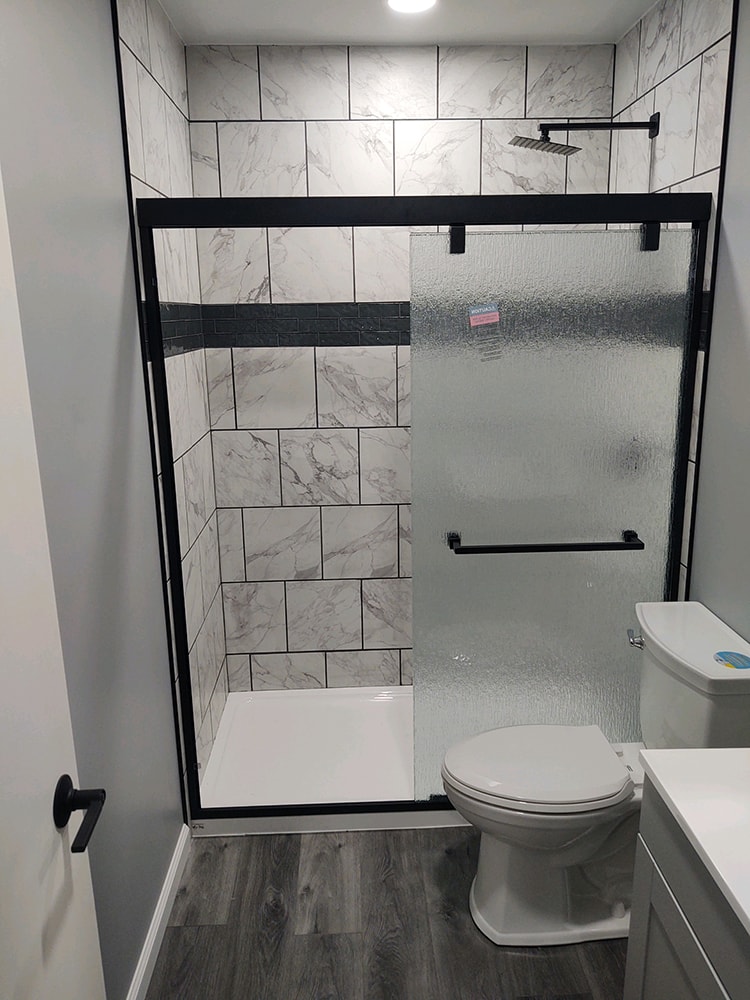 Whole house remodel after — bathroom with marble tile walk-in shower, black framed glass door, rain head, and gray LVP flooring, Fostoria, Ohio